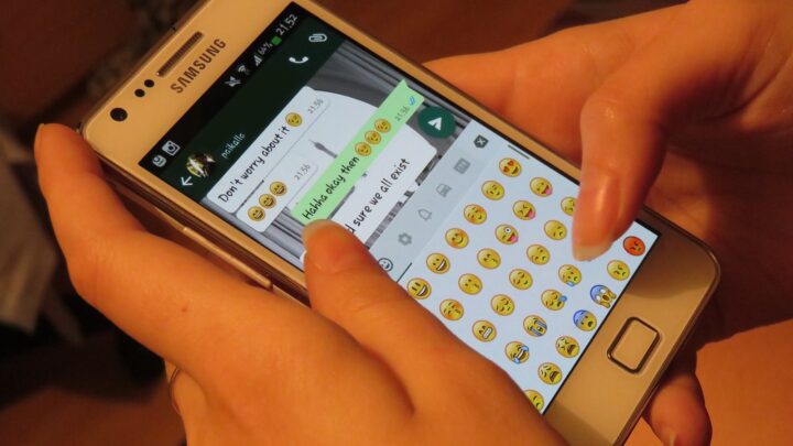 Hands typing on a Samsung smartphone showing a WhatsApp chat with the emoji keyboard open.