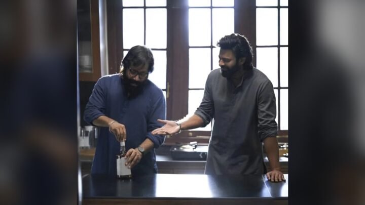 Two men in a kitchen-like room with tall windows: one in a navy shirt and glasses opens a bottle with a plain label, while the other, in a grey kurta, smiles and gestures towards him.